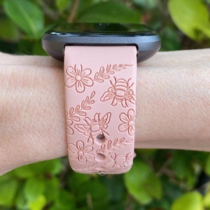 K&ouml;nnte beinhalten: Hellrosa Smartwatch-Armband mit Blumen- und Bienenmuster. Das Armband ist an einer dunkelgrauen Smartwatch befestigt. Das Design umfasst detaillierte Illustrationen von Blumen, Bienen und Bl&auml;ttern. Das Armband besteht aus flexiblem Material.