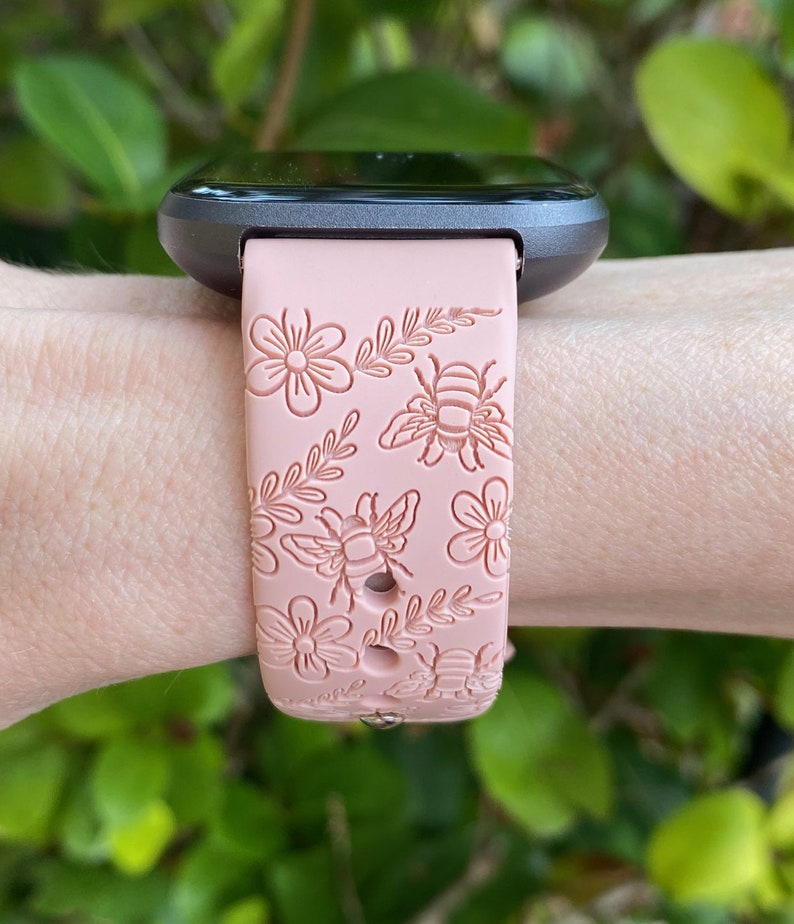 K&ouml;nnte beinhalten: Hellrosa Smartwatch-Armband mit Blumen- und Bienenmuster. Das Armband ist an einer dunkelgrauen Smartwatch befestigt. Das Design umfasst detaillierte Illustrationen von Blumen, Bienen und Bl&auml;ttern. Das Armband besteht aus flexiblem Material.
