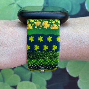 May include: A black smartwatch with a green, gold, and black patterned band featuring shamrocks and glitter. The band is made of silicone and has a secure clasp.