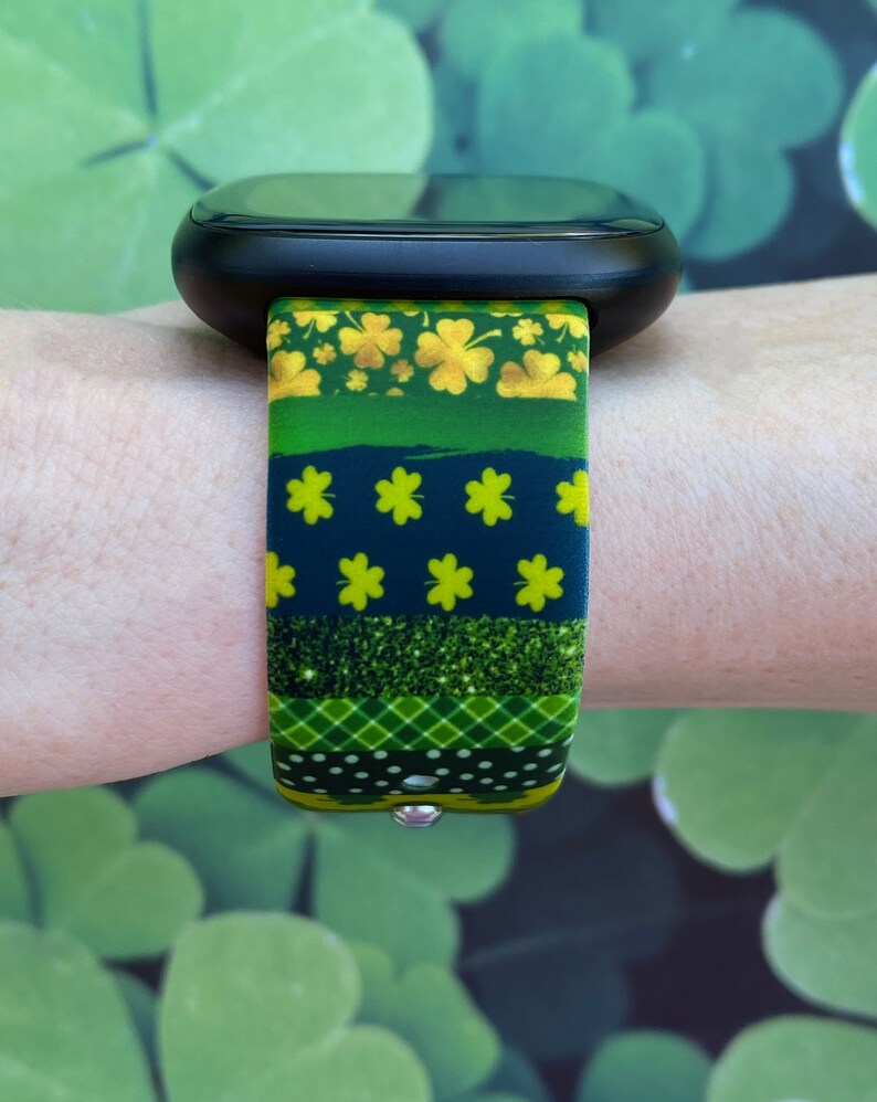 May include: A black smartwatch with a green, gold, and black patterned band featuring shamrocks and glitter. The band is made of silicone and has a secure clasp.