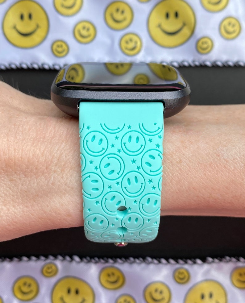 May include: A turquoise smartwatch band with a repeating pattern of smiley faces. The band is made of silicone and has a smooth, matte finish.