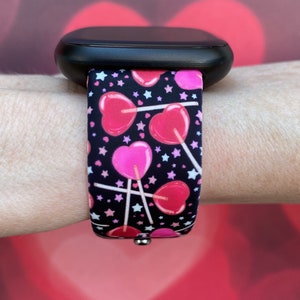 K&ouml;nnte beinhalten: Eine schwarze Smartwatch mit einem Armband im Muster von rosa und roten Herz-Lollies. Das Armband hat wei&szlig;e Sterne auf schwarzem Hintergrund.
