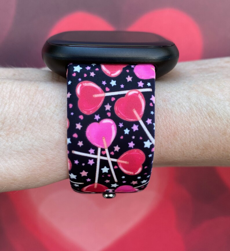 K&ouml;nnte beinhalten: Eine schwarze Smartwatch mit einem Armband im Muster von rosa und roten Herz-Lollies. Das Armband hat wei&szlig;e Sterne auf schwarzem Hintergrund.