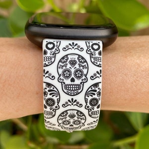 May include: A white and black watch band with a repeating pattern of sugar skulls. The band is made of silicone and is compatible with smartwatches.