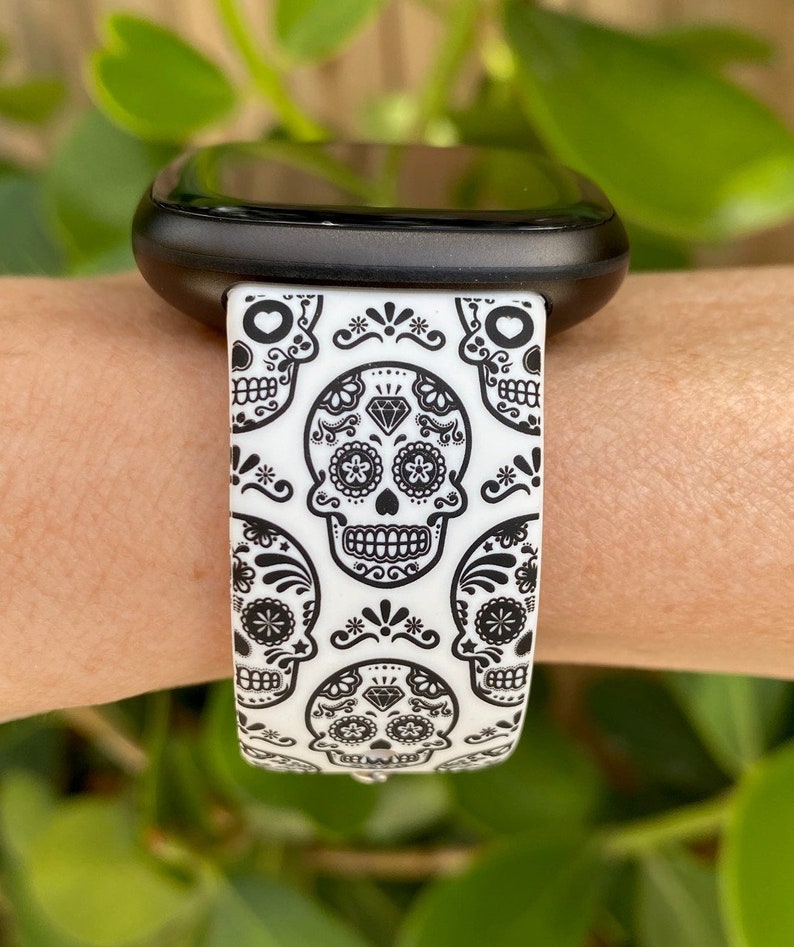 May include: A white and black watch band with a repeating pattern of sugar skulls. The band is made of silicone and is compatible with smartwatches.
