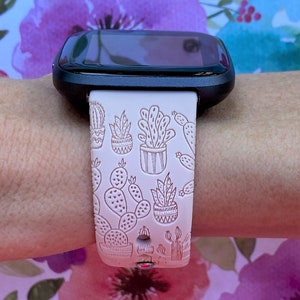 K&ouml;nnte beinhalten: Ein wei&szlig;es Armband f&uuml;r eine Smartwatch mit einem Kaktus-Druckmuster. Das Armband wird am Handgelenk einer Person getragen.