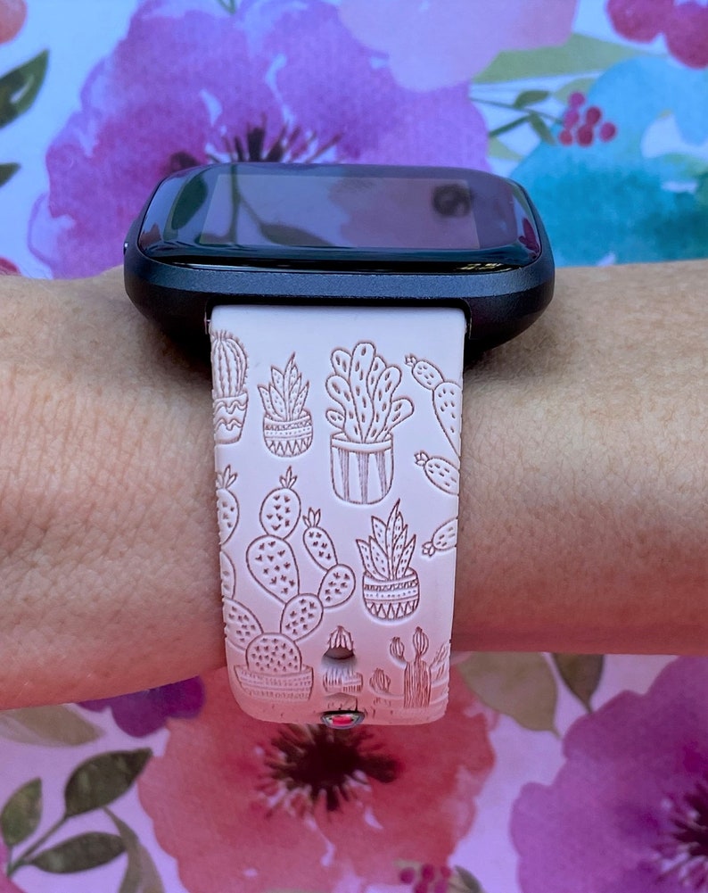 K&ouml;nnte beinhalten: Ein wei&szlig;es Armband f&uuml;r eine Smartwatch mit einem Kaktus-Druckmuster. Das Armband wird am Handgelenk einer Person getragen.