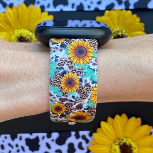 May include: A black smartwatch with a colorful band featuring sunflowers, leopard print, and turquoise accents. The band is secured on a wrist, with yellow sunflowers in the background. The watch band is a vibrant accessory.