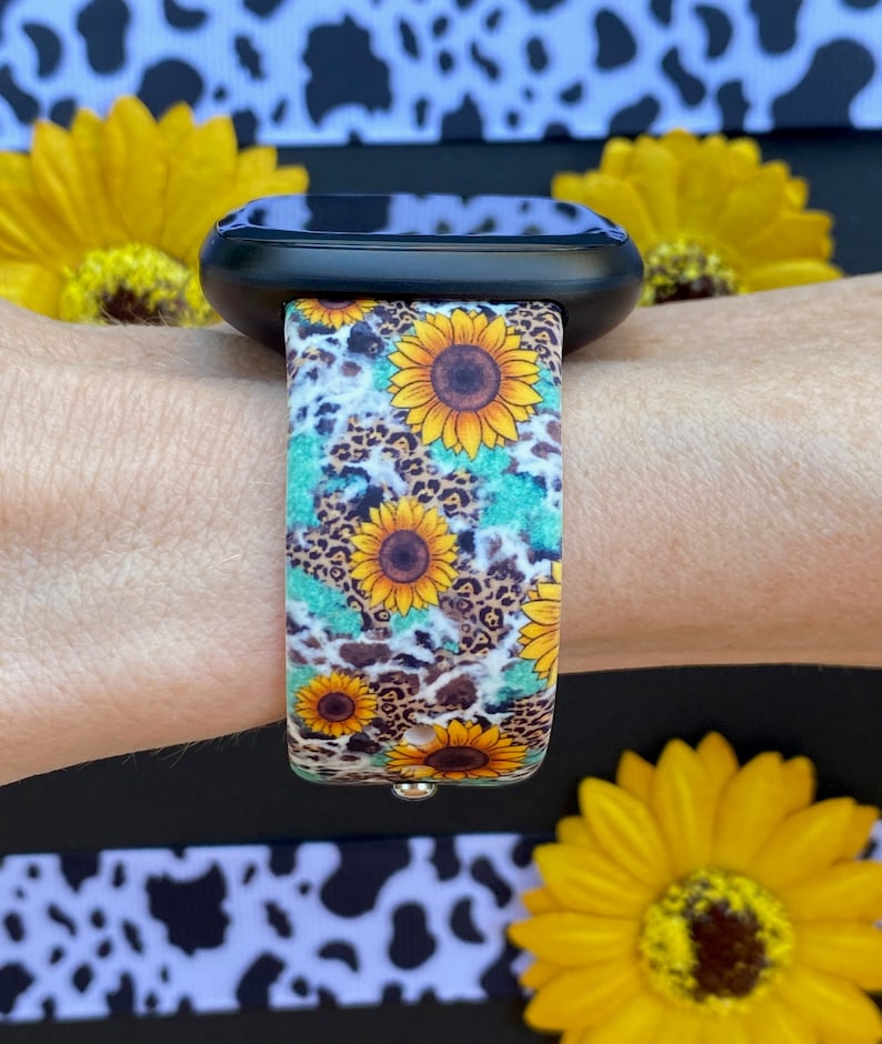 May include: A black smartwatch with a colorful band featuring sunflowers, leopard print, and turquoise accents. The band is secured on a wrist, with yellow sunflowers in the background. The watch band is a vibrant accessory.