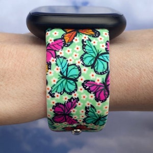 May include: A green watch band with a butterfly pattern. The band has white flowers and butterflies in shades of pink, blue, orange, and yellow.