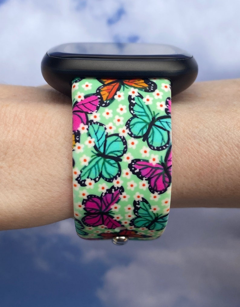 May include: A green watch band with a butterfly pattern. The band has white flowers and butterflies in shades of pink, blue, orange, and yellow.