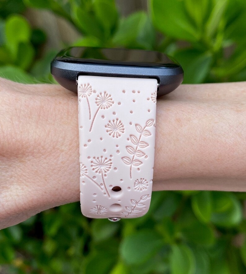May include: A pink silicone watch band with a floral pattern featuring dandelions and leaves. The band is designed for a smartwatch with a black face.