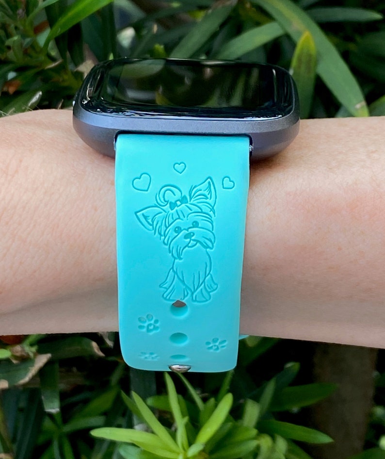 May include: A turquoise blue silicone watch band with a small, white, embossed Yorkshire Terrier design and paw prints. The band is compatible with a smartwatch.