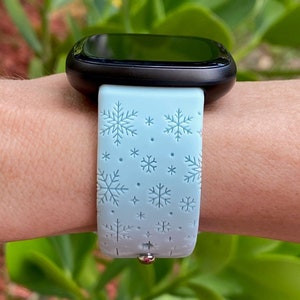 May include: A light blue smartwatch band with a snowflake pattern. The band is made of silicone and has a silver buckle.
