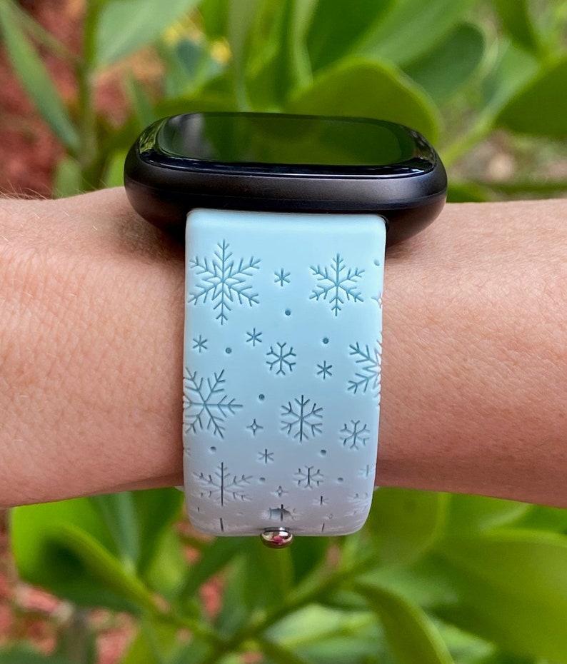 May include: A light blue smartwatch band with a snowflake pattern. The band is made of silicone and has a silver buckle.