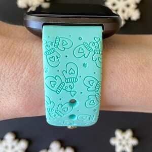 May include: A teal watch band with a repeating pattern of mittens and hearts. The watch band is attached to a black smartwatch. White snowflake decorations are in the background.