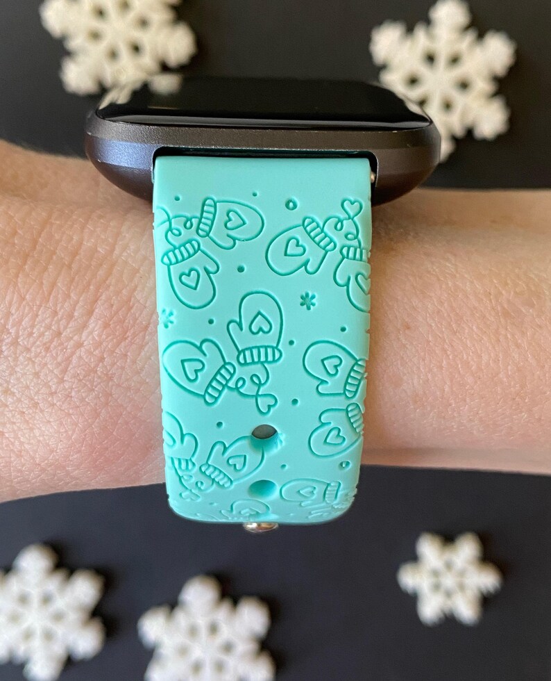 May include: A teal watch band with a repeating pattern of mittens and hearts. The watch band is attached to a black smartwatch. White snowflake decorations are in the background.