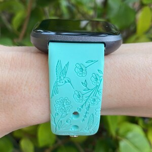 May include: A turquoise smartwatch band with a floral and hummingbird design. The band is made of silicone and has a black watch face.