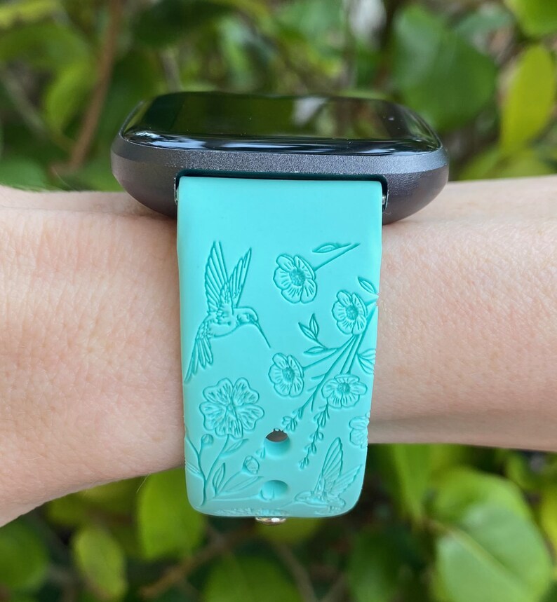 May include: A turquoise smartwatch band with a floral and hummingbird design. The band is made of silicone and has a black watch face.