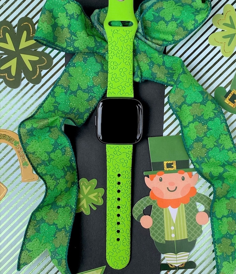 May include: A green silicone watch band with a shamrock pattern. The band is on a black surface with a green and gold ribbon with a shamrock pattern.