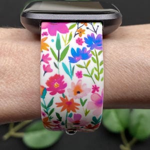 Pu&ograve; includere: Un cinturino per orologio con motivo floreale con fiori rosa, arancioni e blu su sfondo bianco. Il cinturino &egrave; attaccato a uno smartwatch nero. Il design comprende foglie e steli verdi.