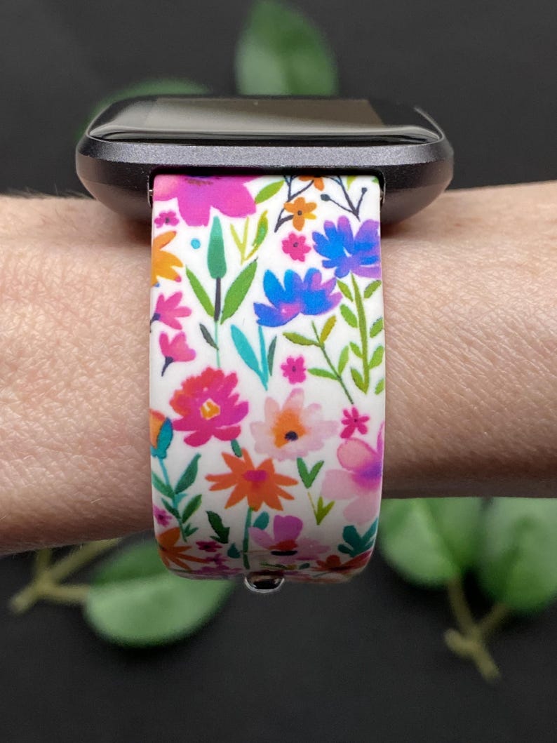 Pu&ograve; includere: Un cinturino per orologio con motivo floreale con fiori rosa, arancioni e blu su sfondo bianco. Il cinturino &egrave; attaccato a uno smartwatch nero. Il design comprende foglie e steli verdi.