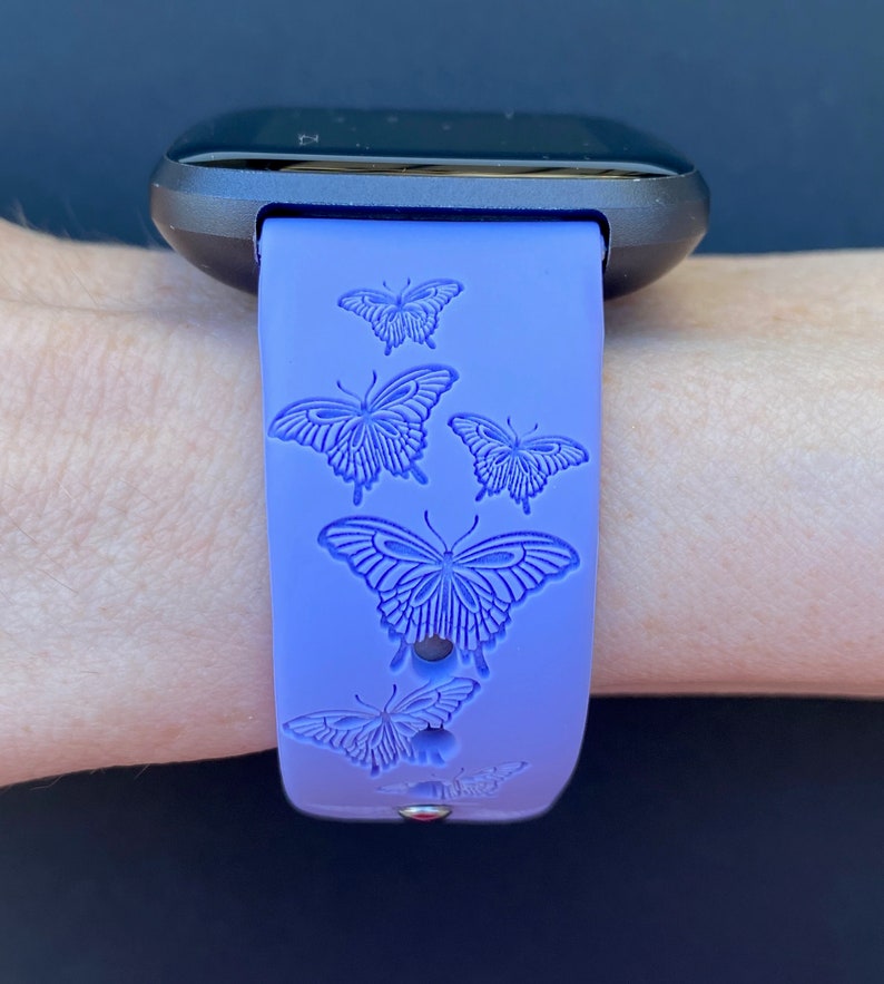 May include: A purple silicone watch band with a black watch face. The band has a repeating pattern of embossed butterflies.