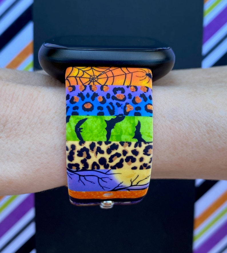 May include: A colorful watch band with a Halloween theme. The band features a spiderweb, bats, leopard print, and a moon with branches. The band is made of silicone and is compatible with most smartwatches.