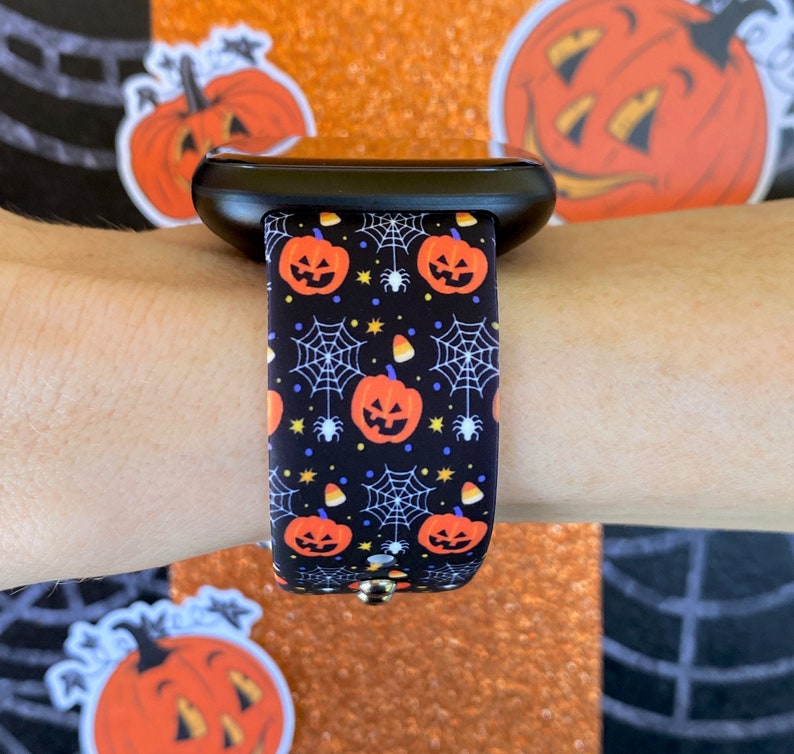 May include: A black smartwatch with a Halloween-themed band. The band features a repeating pattern of orange pumpkins, spiderwebs, and candy corn on a black background.