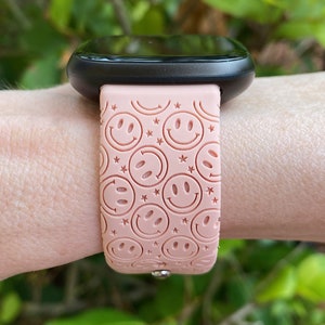 May include: A pink smartwatch band with a pattern of smiling faces and stars. The band is made of silicone and has a metal clasp.