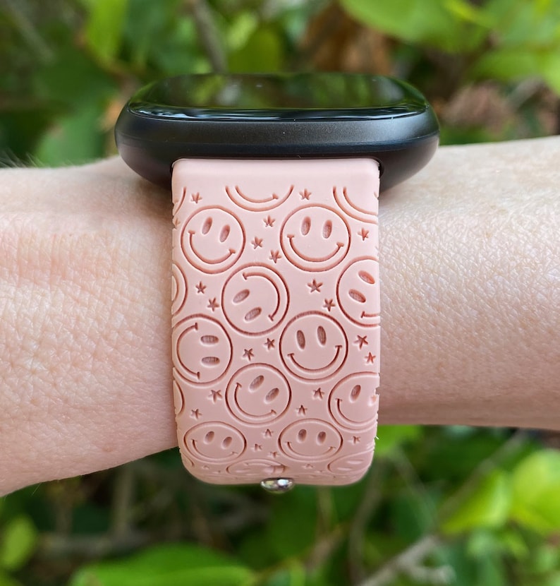 May include: A pink smartwatch band with a pattern of smiling faces and stars. The band is made of silicone and has a metal clasp.