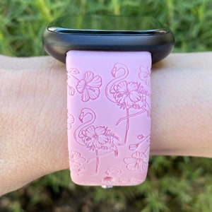 May include: Pink watch band with a flamingo and floral design. The band is attached to a black smartwatch. The flamingo and floral pattern is embossed on the pink band.