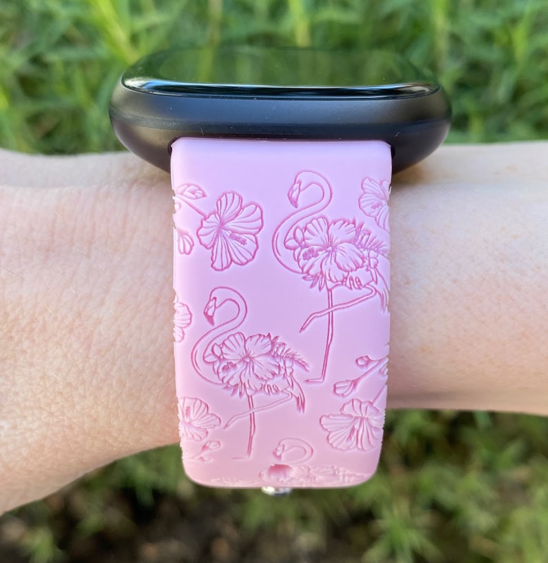 May include: Pink watch band with a flamingo and floral design. The band is attached to a black smartwatch. The flamingo and floral pattern is embossed on the pink band.