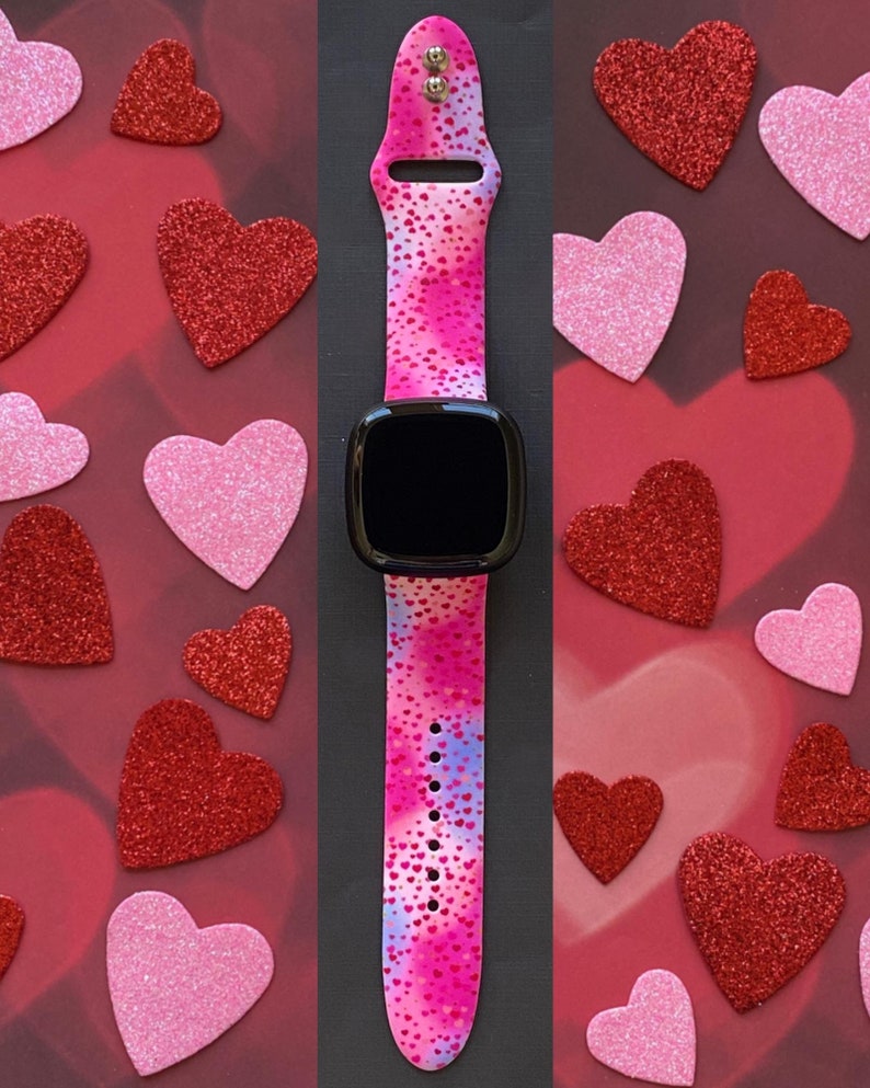 May include: A pink and blue smartwatch band with a white background and small pink hearts printed on it. The band is made of silicone and has a black watch face.