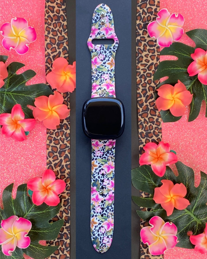 May include: A black smartwatch with a floral and leopard print silicone strap. The strap has pink, white, and green flowers on a white background with a leopard print overlay.
