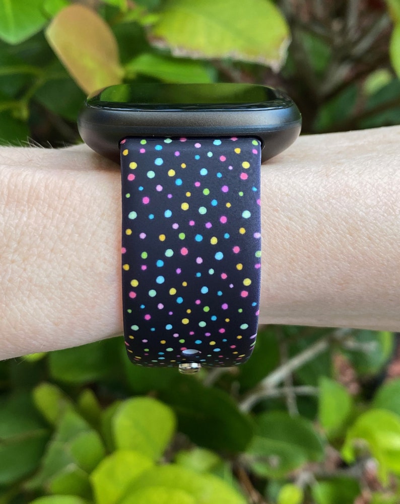 May include: A black smartwatch with a colorful polka dot patterned band. The band has a variety of colors including pink, blue, green, and yellow.