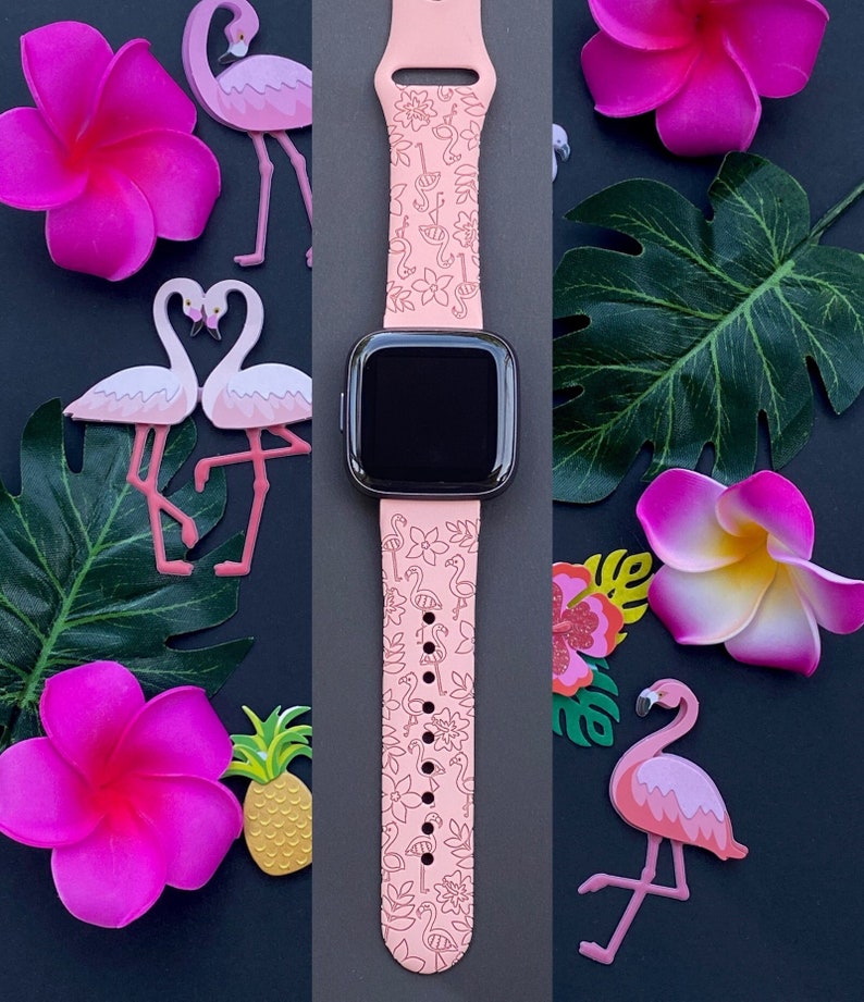 May include: A pink Apple Watch band with a flamingo and floral pattern. The band is shown on a black background with pink flowers and a pineapple.