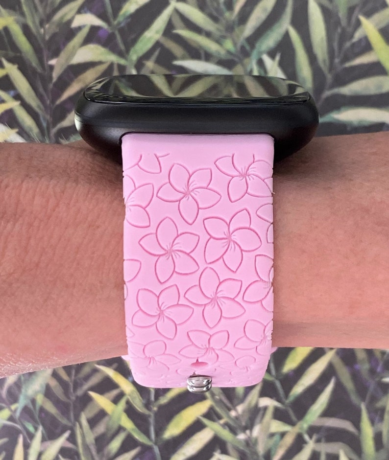 May include: A pink silicone watch band with a floral pattern. The band is designed for a smartwatch and has a silver buckle.