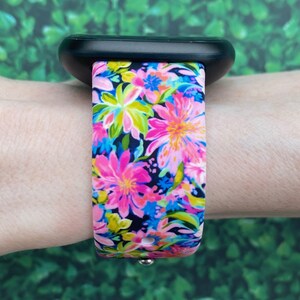 May include: A black smartwatch with a pink and blue floral patterned band. The band is made of silicone and has a black background with pink, blue, yellow, and green flowers.