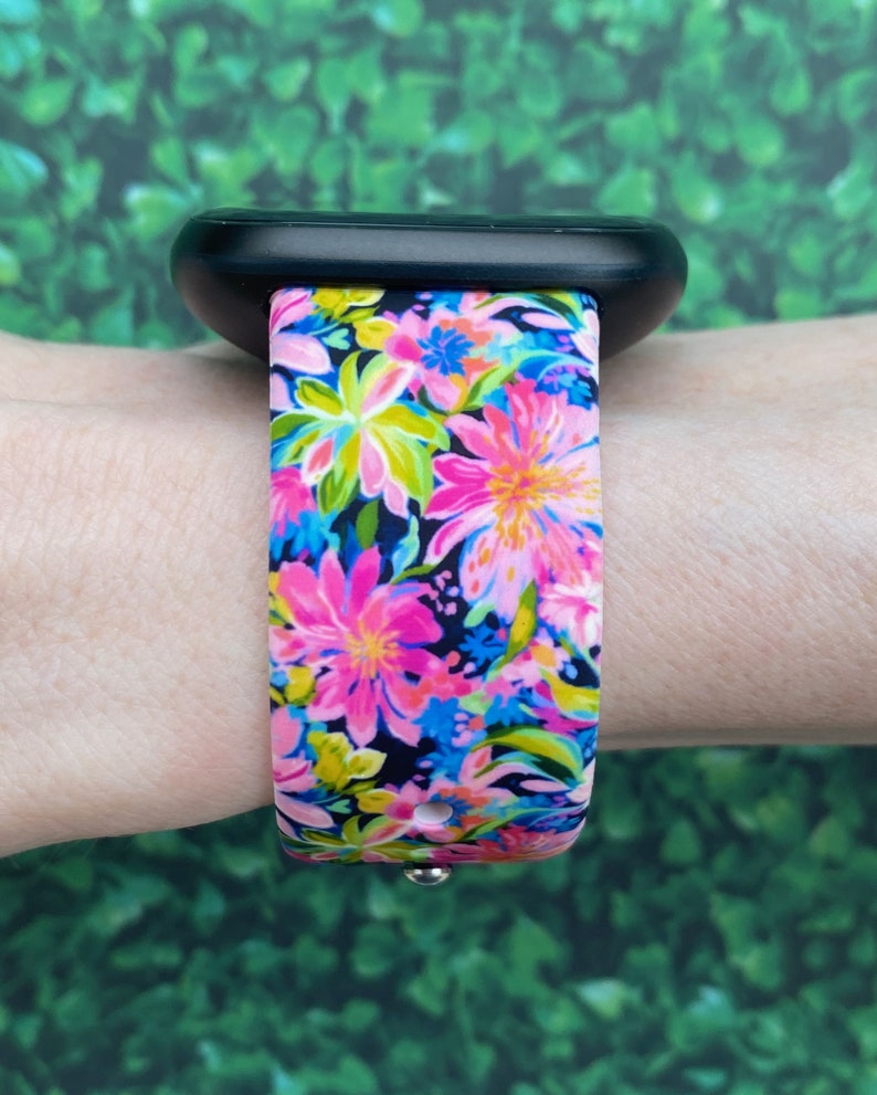 May include: A black smartwatch with a pink and blue floral patterned band. The band is made of silicone and has a black background with pink, blue, yellow, and green flowers.