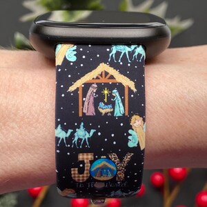 Pu&ograve; includere: Cinturino nero per orologio con un design a scena della nativit&agrave; natalizia. Il cinturino presenta illustrazioni della Sacra Famiglia, angeli, cammelli e il testo "Joy To The World". Il cinturino &egrave; attaccato a uno smartwatch nero.