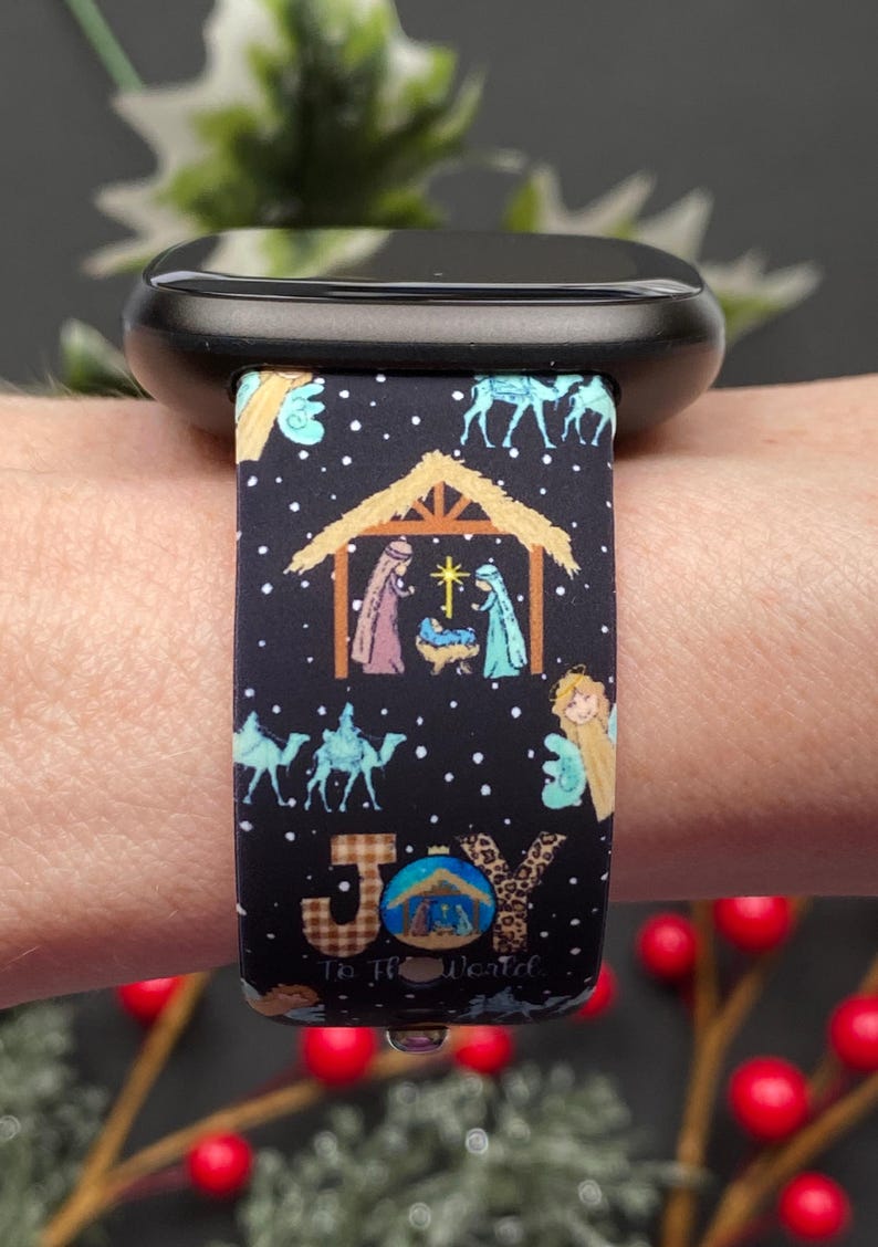 Pu&ograve; includere: Cinturino nero per orologio con un design a scena della nativit&agrave; natalizia. Il cinturino presenta illustrazioni della Sacra Famiglia, angeli, cammelli e il testo "Joy To The World". Il cinturino &egrave; attaccato a uno smartwatch nero.