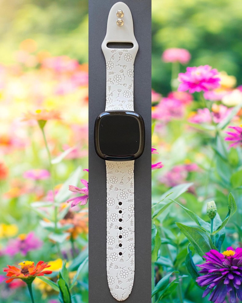 K&ouml;nnte beinhalten: Wei&szlig;es Silikon-Armband mit Blumenmuster. Das Armband ist an einer schwarzen Smartwatch mit rundem Zifferblatt befestigt.