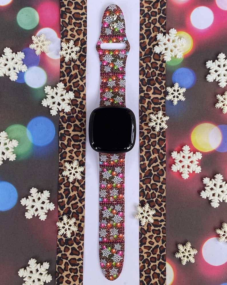 May include: A black smartwatch with a colourful Christmas lights and snowflakes patterned band. The band is on a brown leopard print background.