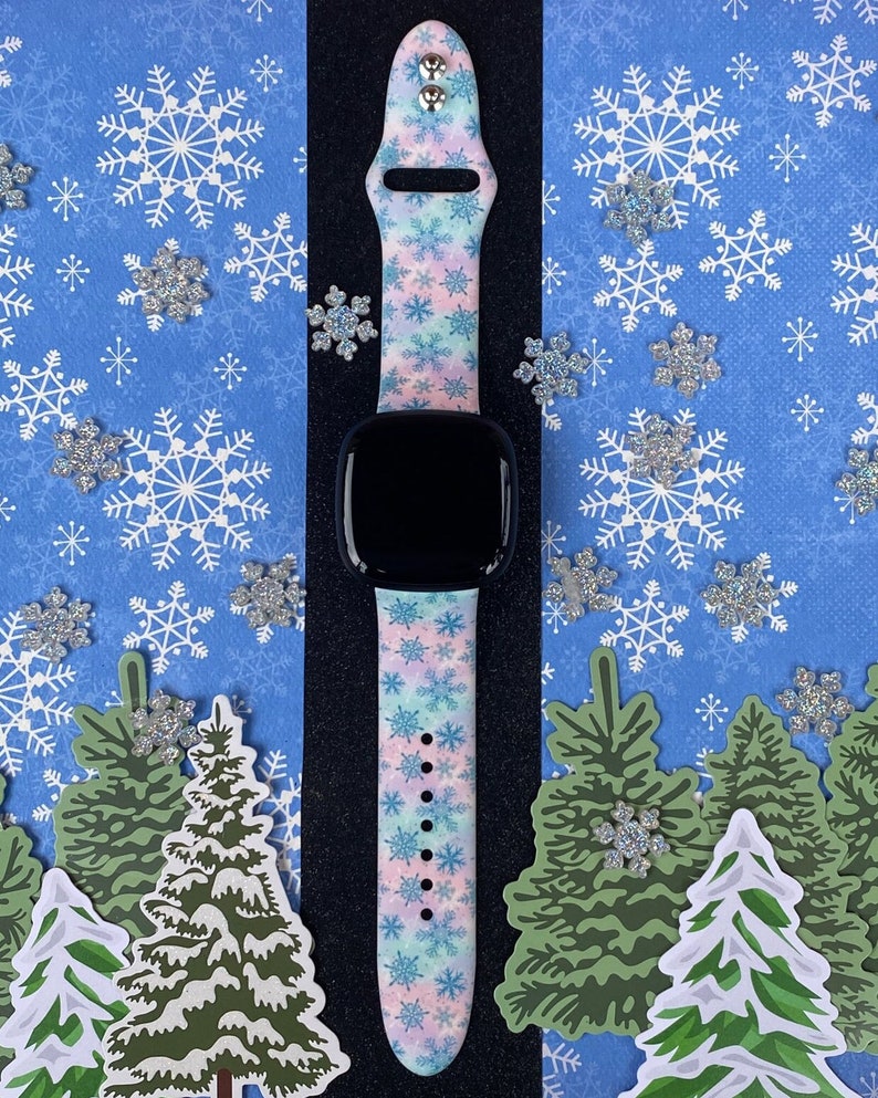 May include: A pink, blue, and white Apple Watch band with a snowflake pattern. The band is on a black background with a white snowflake pattern.