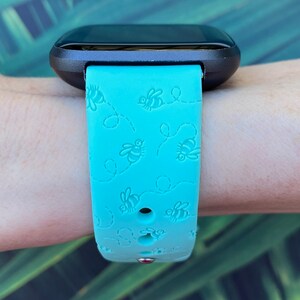 May include: A turquoise blue fitness tracker band with a black watch face. The band has a repeating pattern of bees and dotted lines.