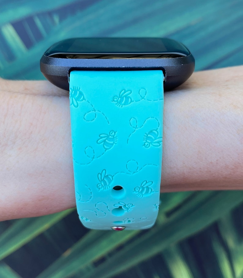 May include: A turquoise blue fitness tracker band with a black watch face. The band has a repeating pattern of bees and dotted lines.