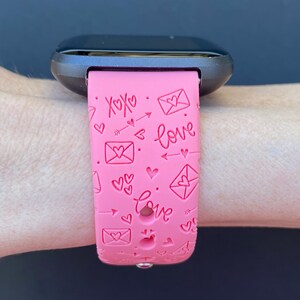May include: Pink smartwatch band with a pattern of hearts, arrows, and envelopes with the words "xoxo" and "love" embossed on it.