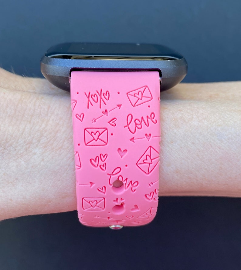 May include: Pink smartwatch band with a pattern of hearts, arrows, and envelopes with the words "xoxo" and "love" embossed on it.
