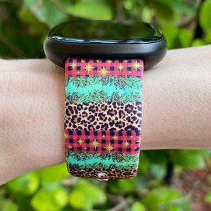 Op de afbeelding: Een zwarte smartwatch met een kleurrijk kerstbandje. Het bandje heeft een rood-zwart ruitpatroon, groene dennentakken, gouden sterren en een luipaardprint.